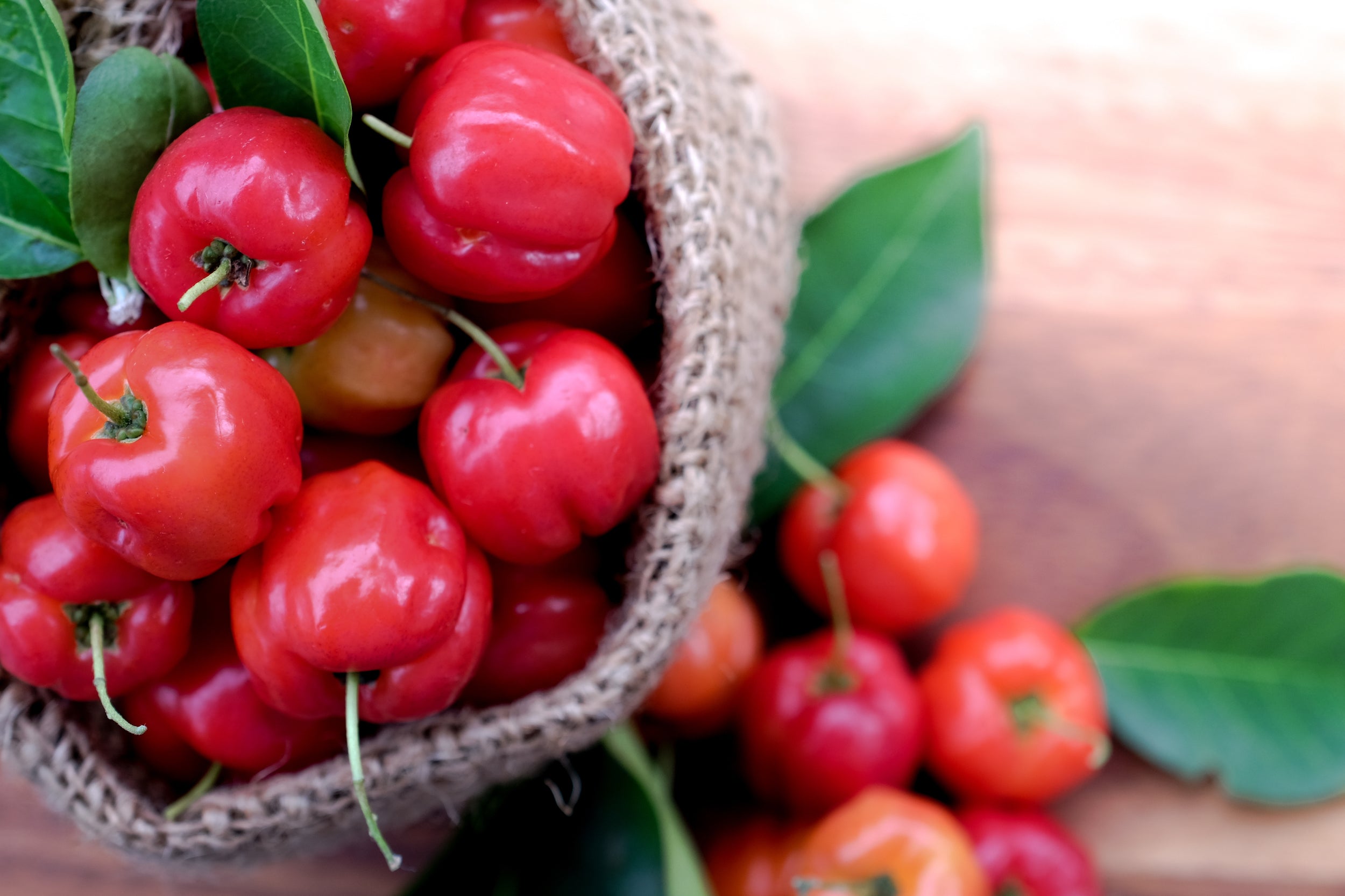 A basket full of bright red acerola cherries overflowing onto a wooden surface